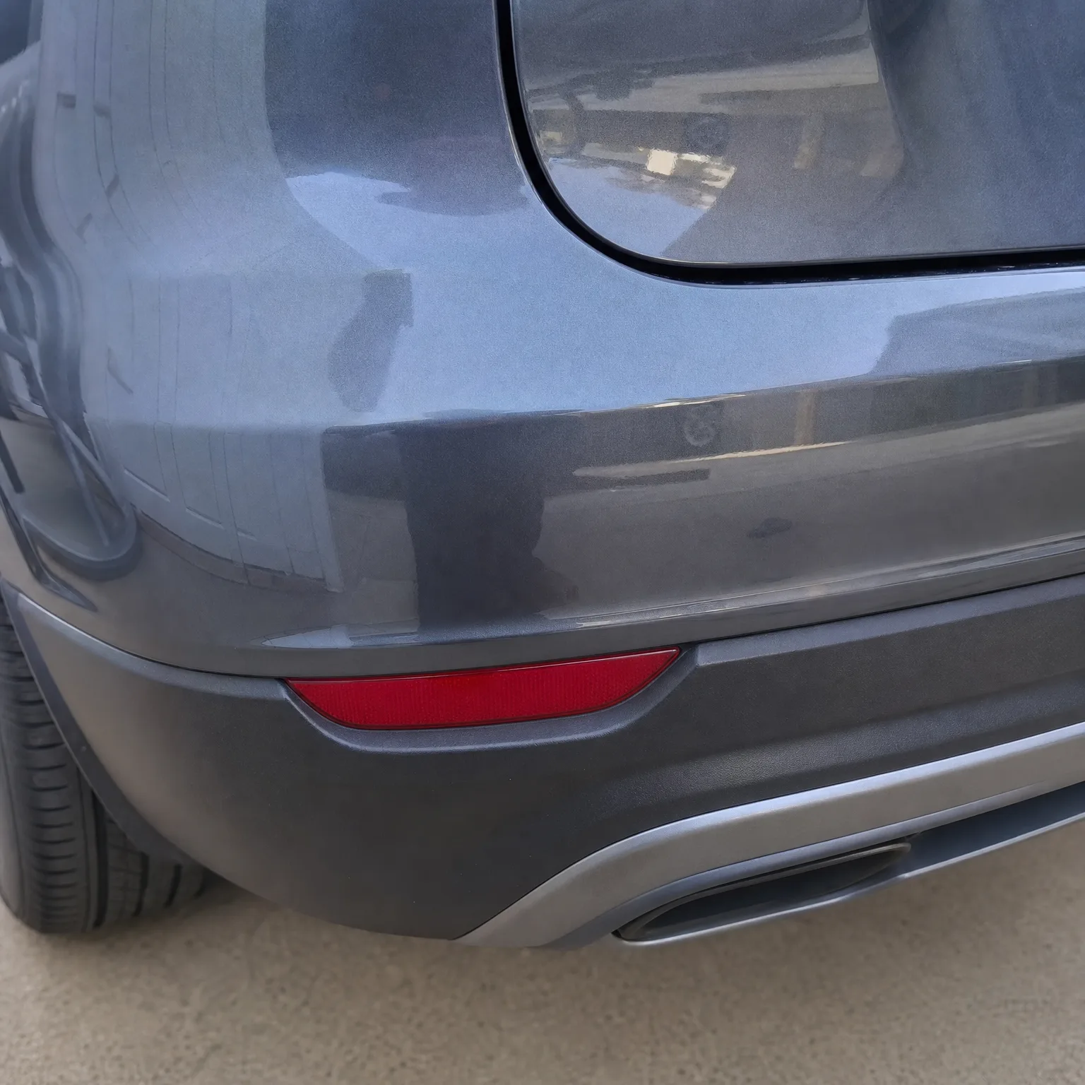 Close-up of the rear corner of a gray car showing the bumper with a red reflector, part of the rear window, a tire, and a chrome exhaust tip above pale pavement.