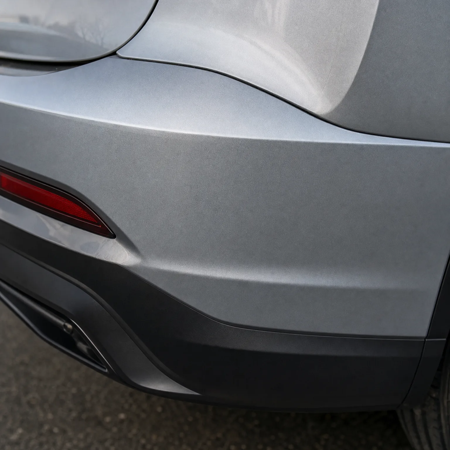 Close-up of the rear corner of a silver-grey car, showing the bumper with a red reflector, dark lower trim, exhaust outlet and part of a tyre on asphalt.