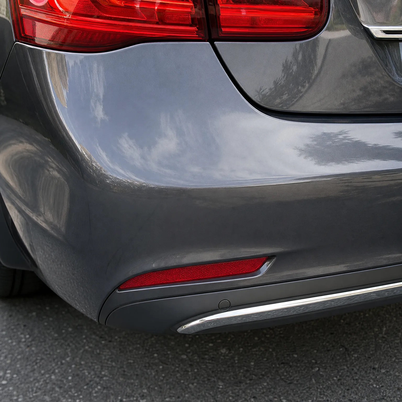 Close-up of the rear corner of a gray car, showing the taillight, bumper with a red reflector strip, chrome trim and a parking sensor, above asphalt pavement.