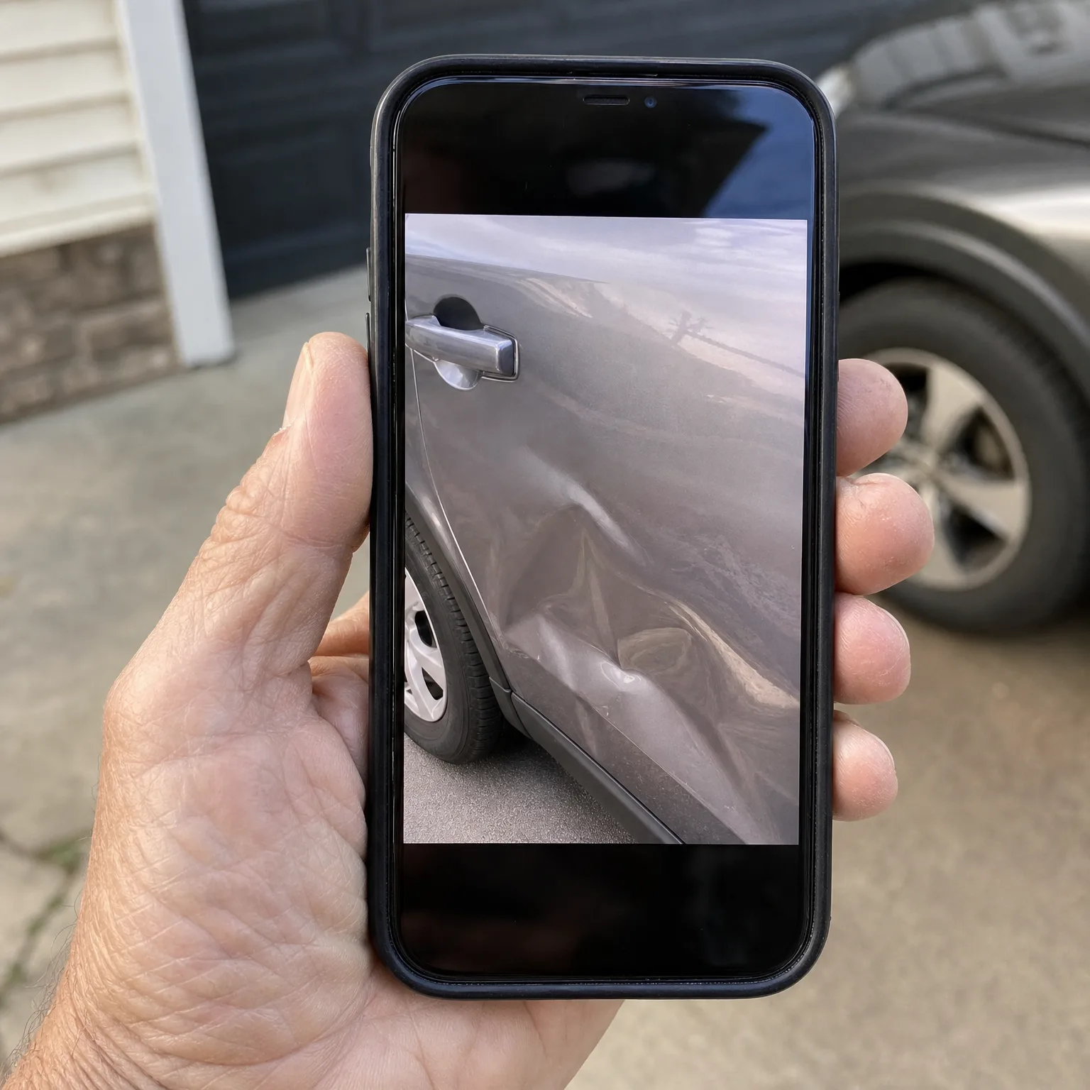 A hand holds a smartphone in a black case displaying a photo of a dented gray car door panel, with a parked vehicle and driveway blurred in the background.