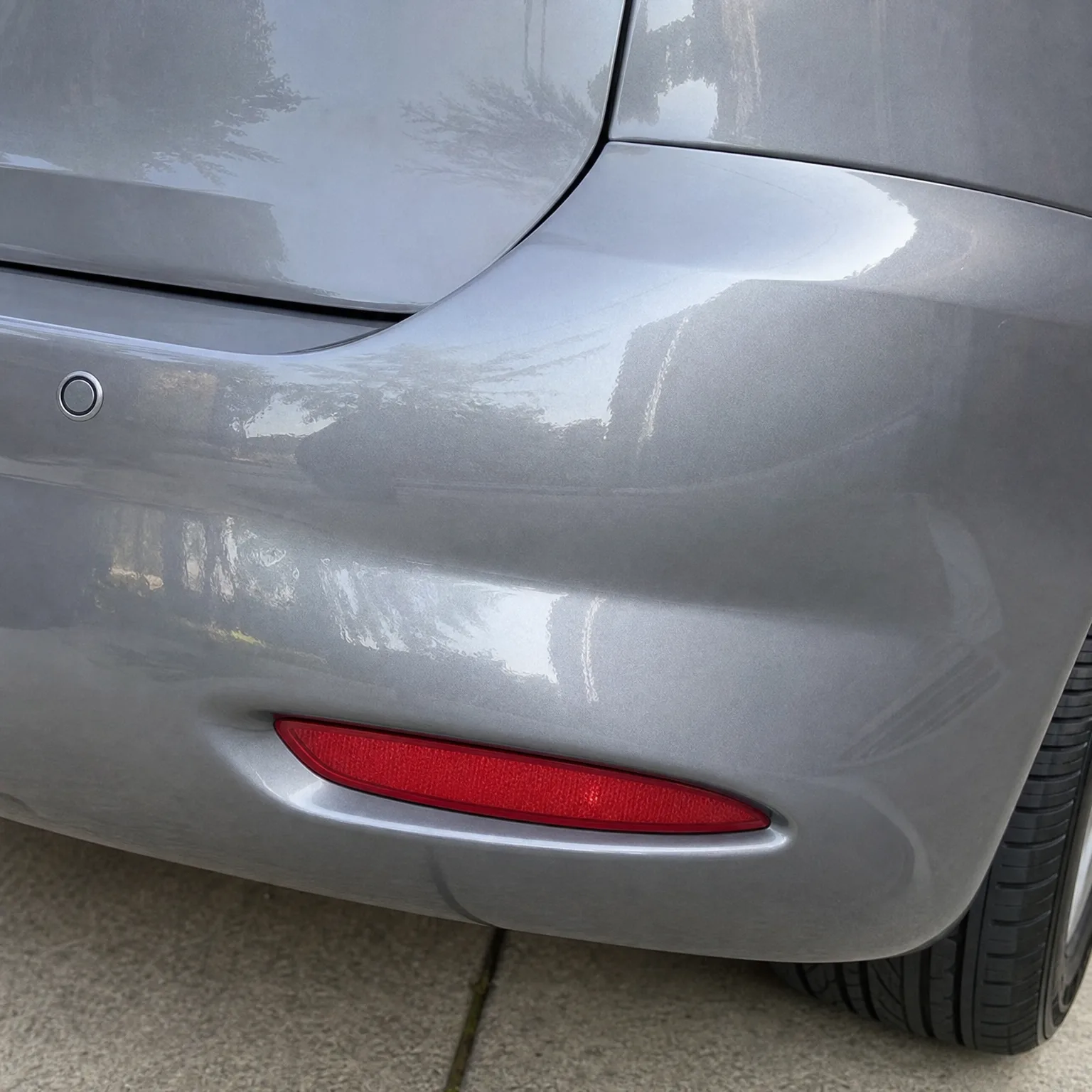 Close-up of the rear corner of a silver-grey car showing a red bumper reflector, a round parking sensor, and part of the tyre above a concrete surface.