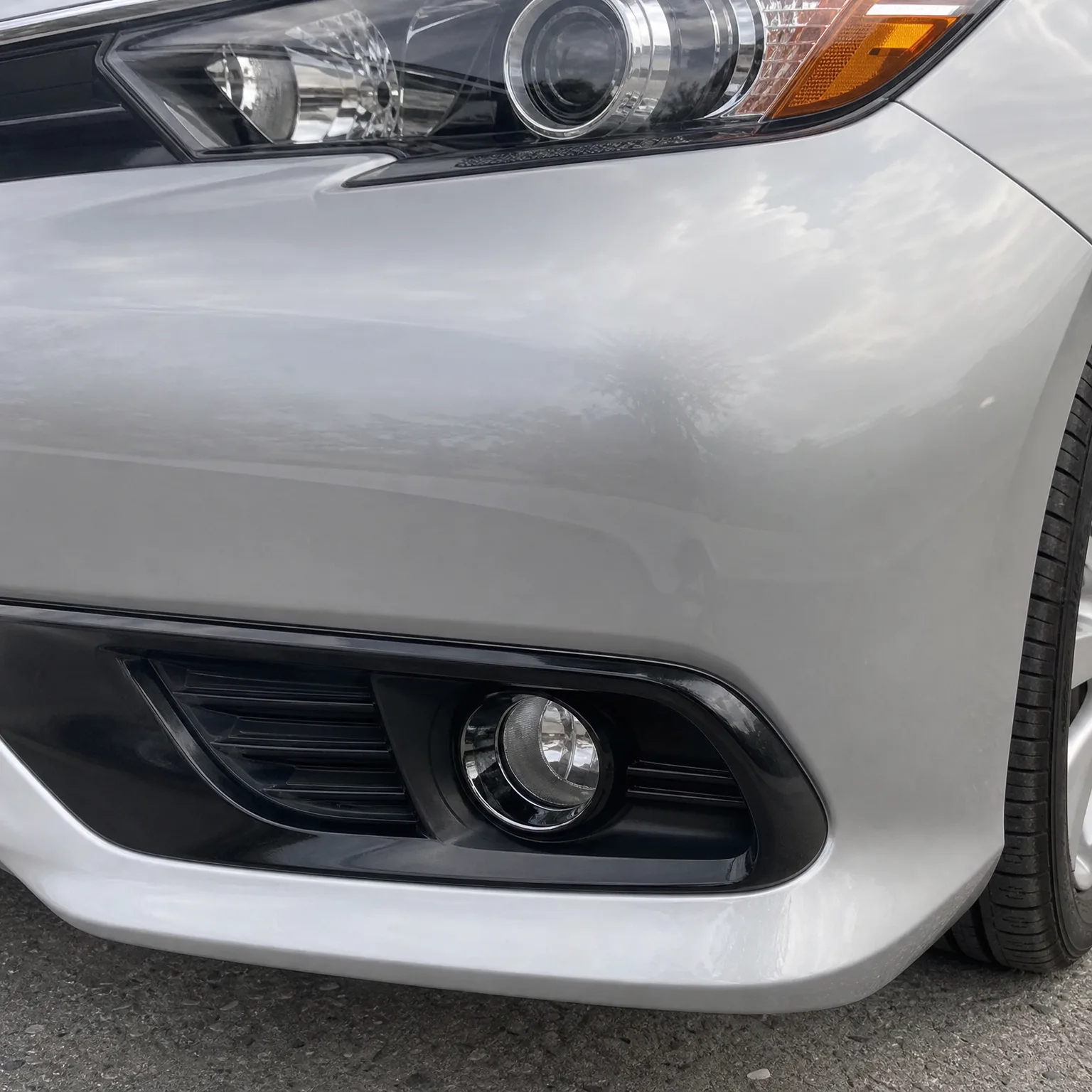 Close-up of the front left corner of a silver car, showing the headlight edge, bumper, black fog light bezel with grille vent, and part of the front tire on pavement.
