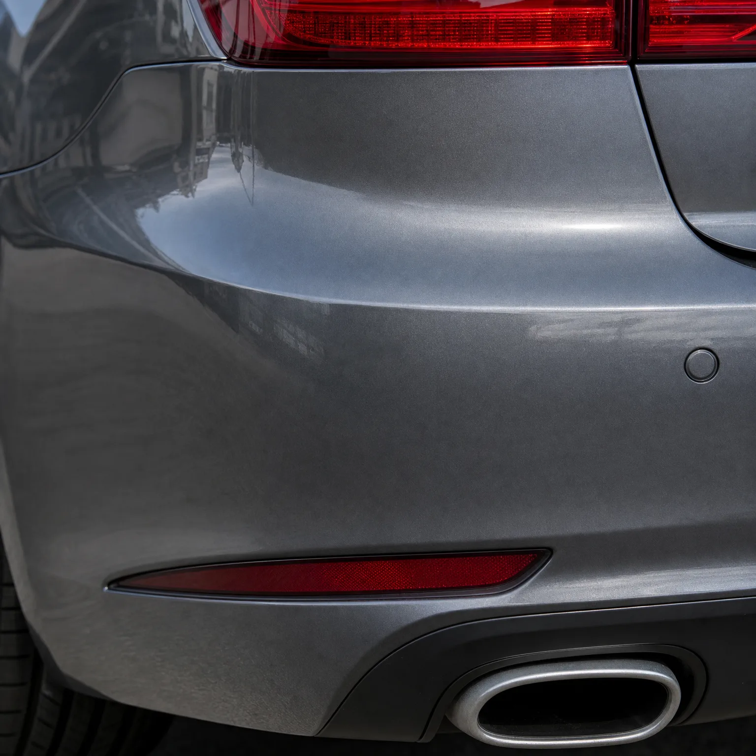 Close-up of the rear corner of a gray car showing part of the red taillight, a red bumper reflector, a round parking sensor, a chrome-trimmed exhaust tip, and the edge of a tire.