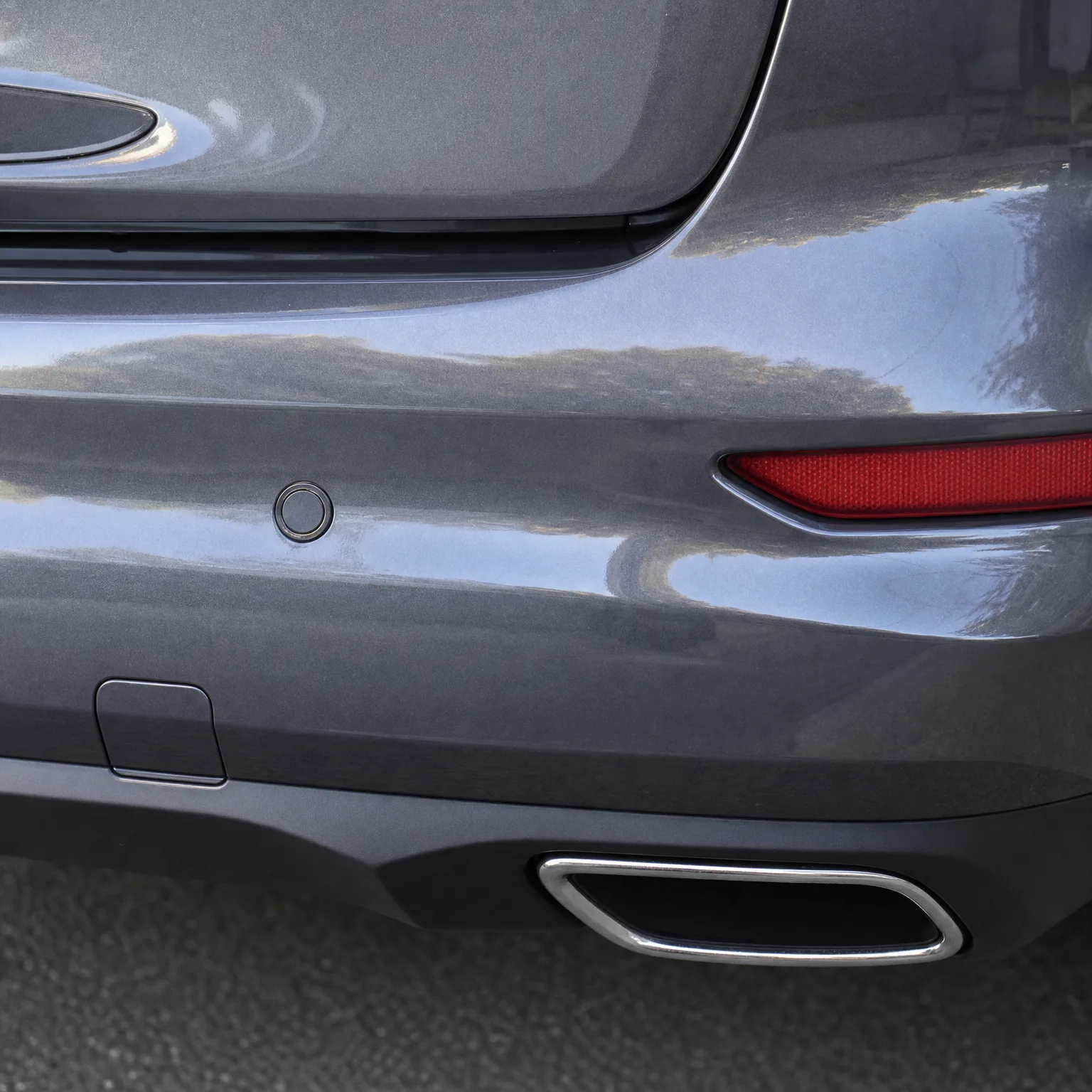 Close-up of the rear corner of a gray car showing a red reflector, a parking sensor, a bumper access cover, and a chrome-trimmed rectangular exhaust outlet above pavement.