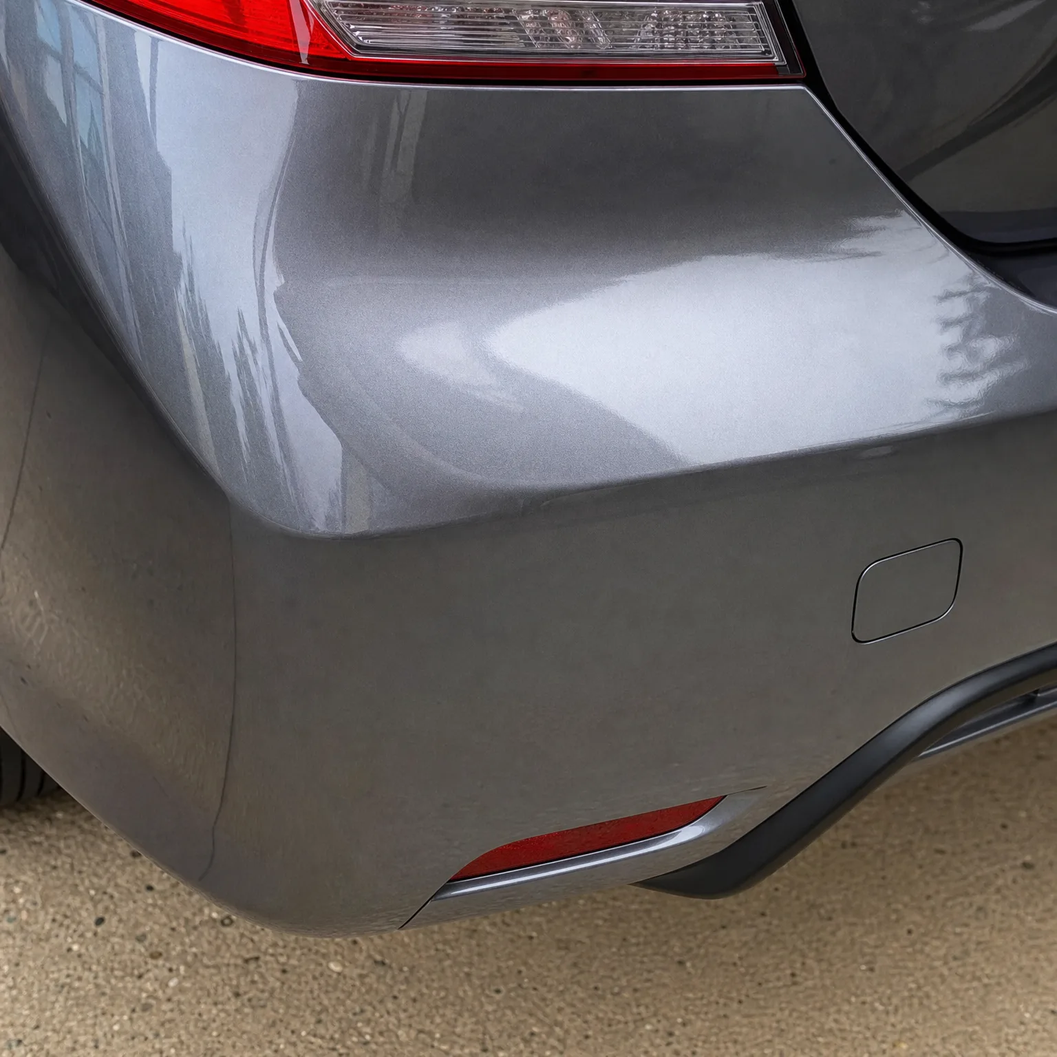 Close-up of the rear corner of a gray car, showing the bumper, red reflector, taillight edge and tire above a concrete surface.