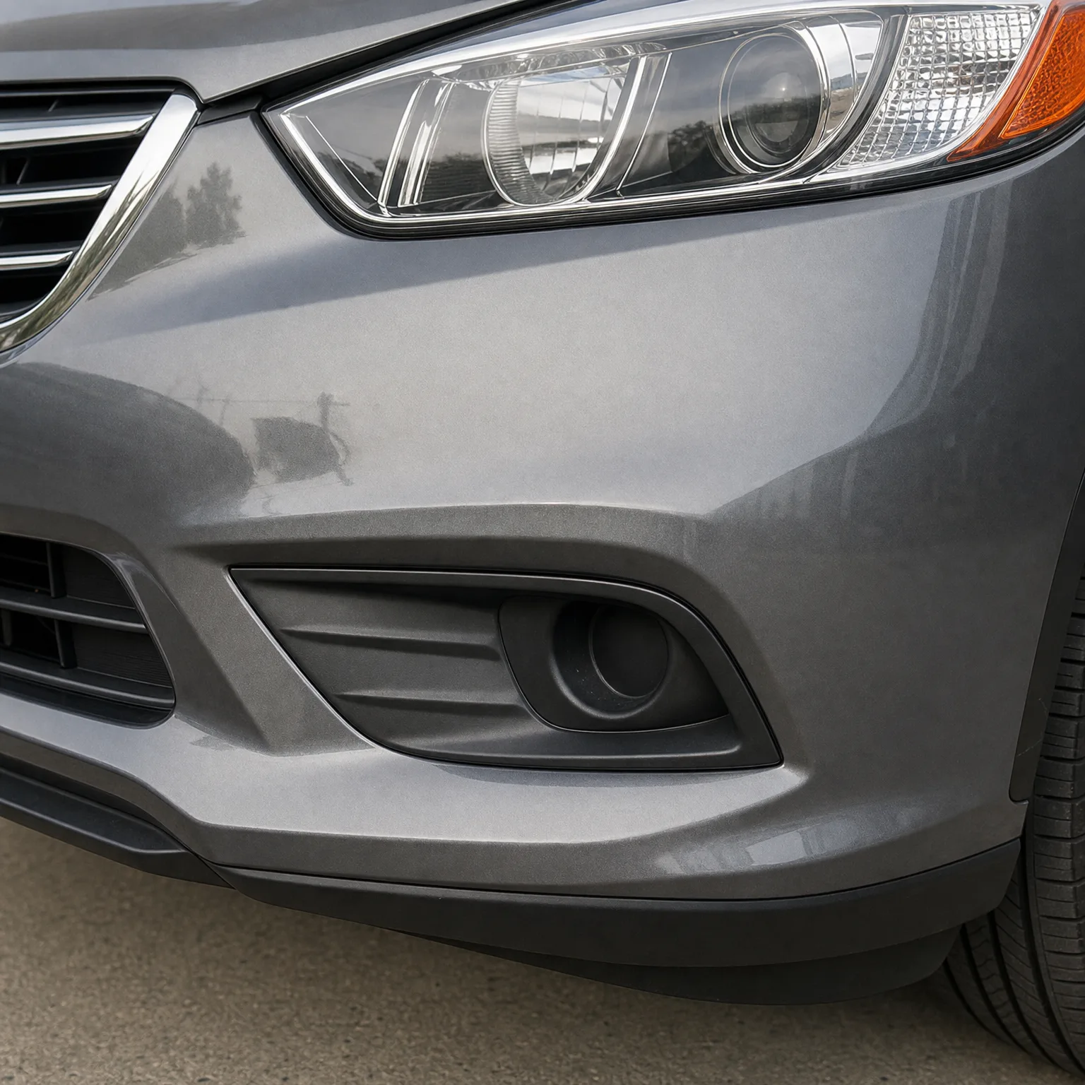 Close-up of the front corner of a gray car showing the headlight, bumper, fog light housing and part of the front tire above pavement.