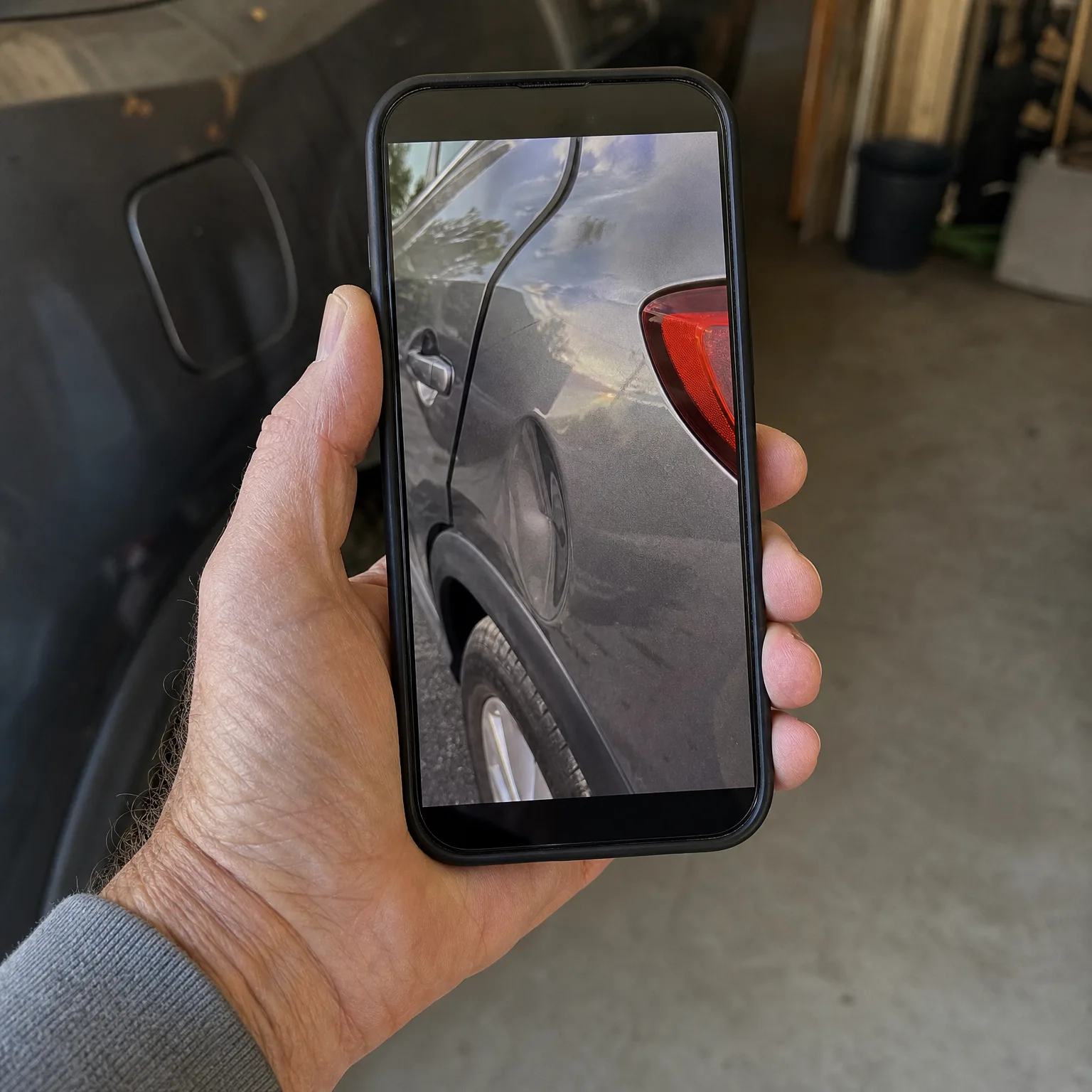 A hand holds a smartphone displaying a photo of a dark car's rear quarter panel and taillight, with a dark vehicle and concrete garage floor in the background.