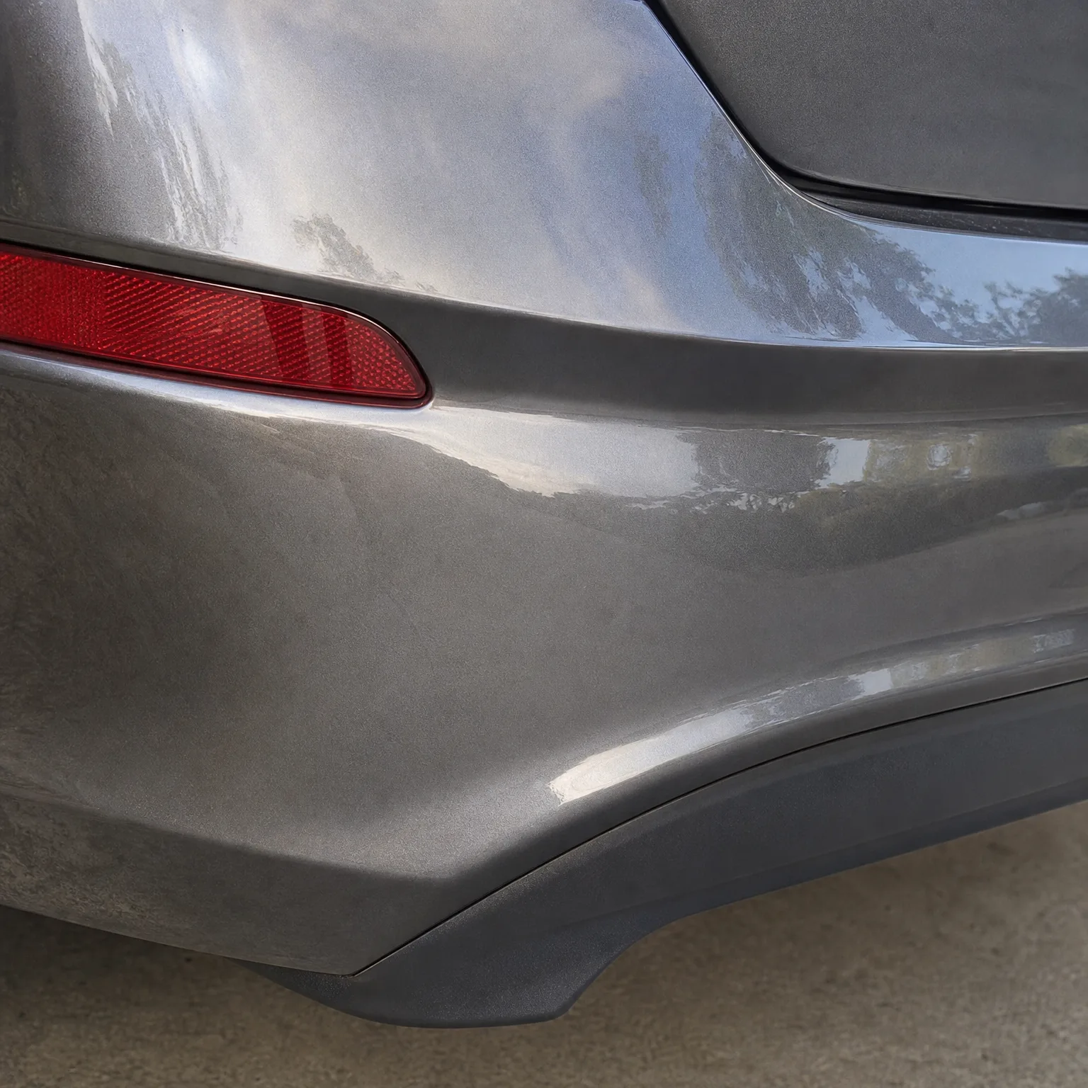 Close-up of the rear corner of a gray car's bumper with a red reflector and a black lower trim piece, above a concrete surface.