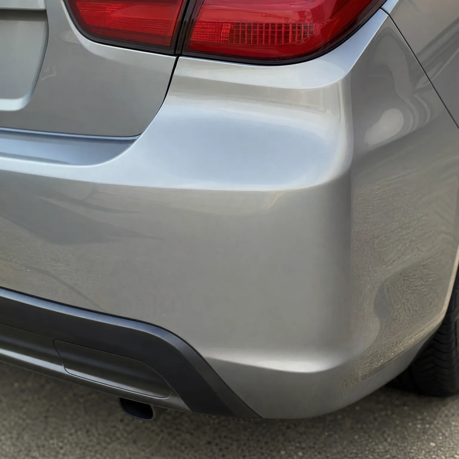 Close-up of the rear corner of a silver car showing the bumper, part of a red taillight, lower black trim with exhaust tip, and a tire above asphalt pavement.