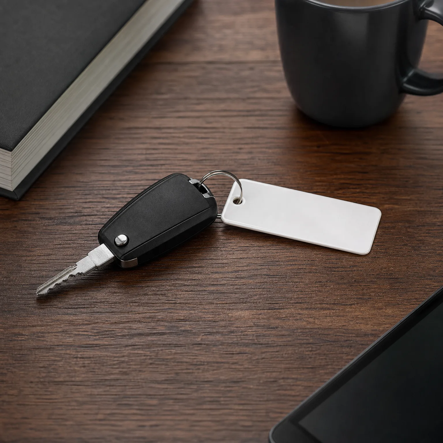 A black flip car key with a blank white rectangular keychain tag on a dark wood surface, with a hardcover book, a dark mug, and the corner of a tablet nearby.
