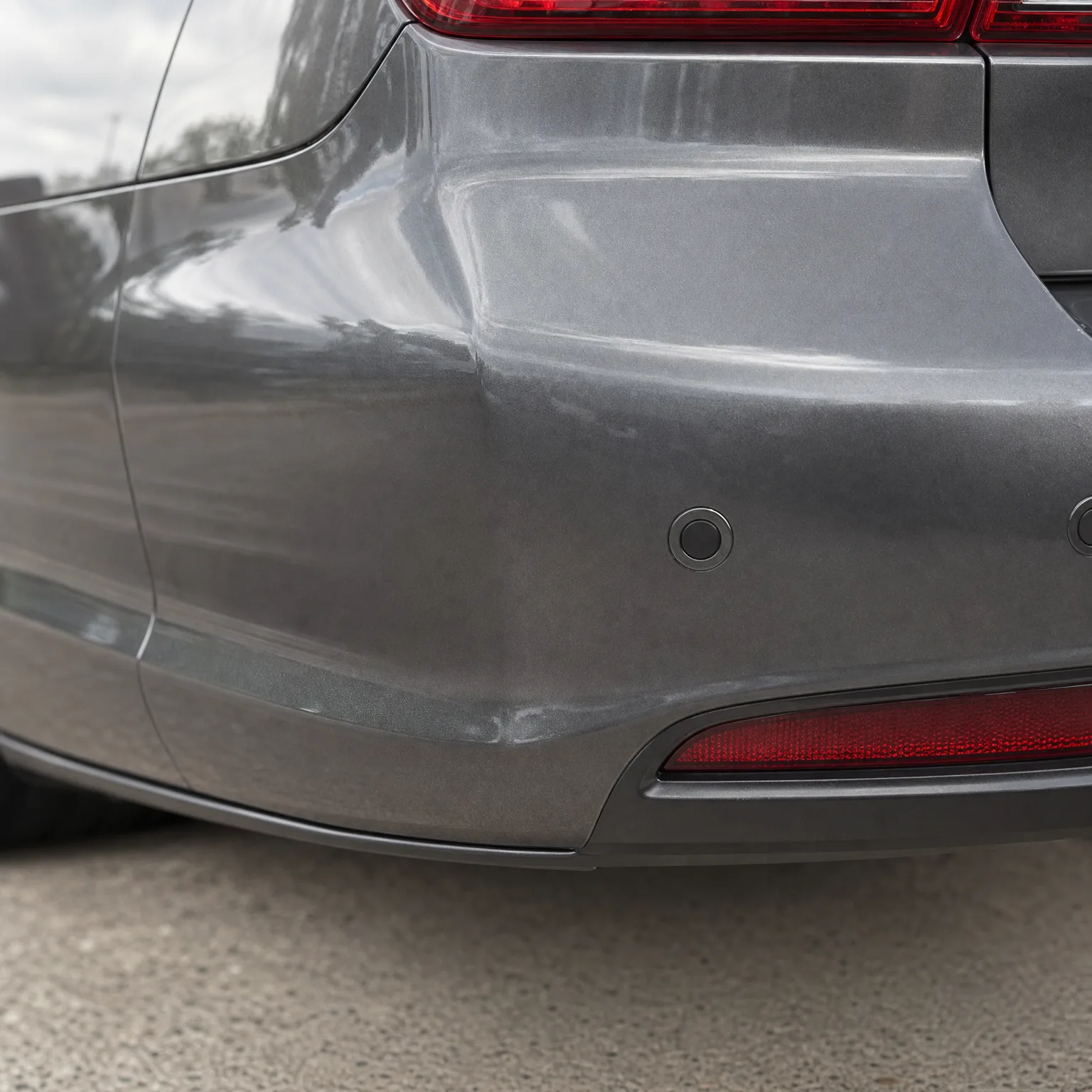 Close-up of the rear corner of a gray car showing the bumper with a parking sensor, red reflector, part of a taillight, and pavement below.