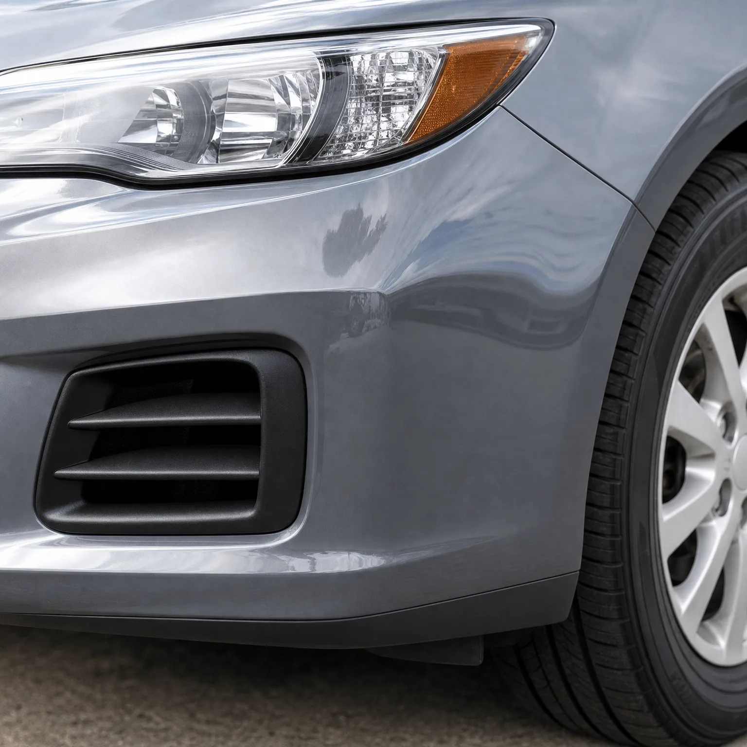 Close-up of the front left corner of a gray car showing the headlight, bumper with a black louvered fog light cover, front tire and alloy wheel, on pavement.