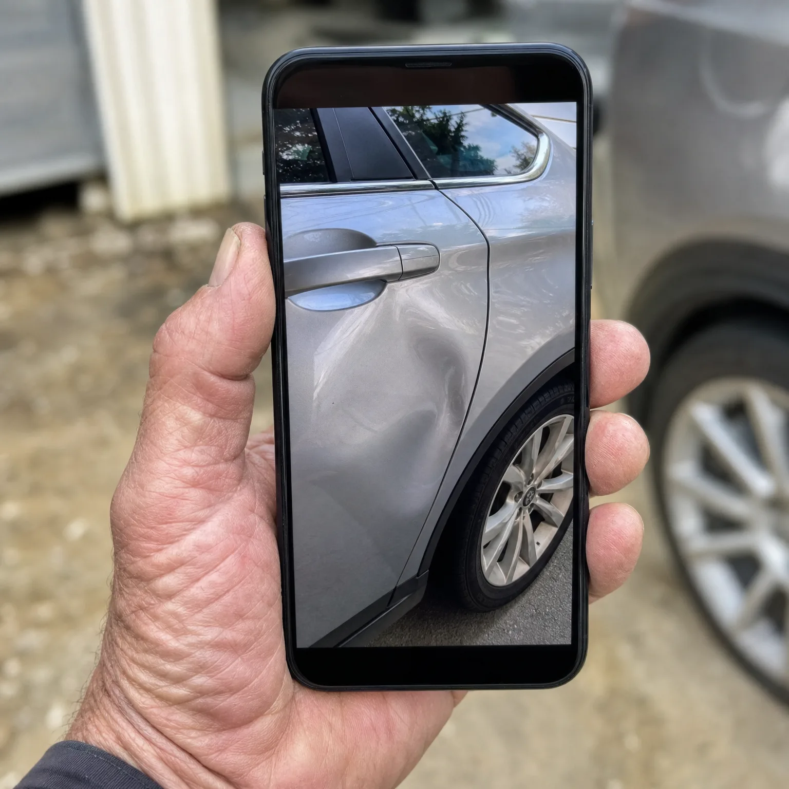 A hand holds a smartphone displaying a close-up photo of a dent on a silver car's rear door and fender, with a parked vehicle and gravel ground blurred in the background.