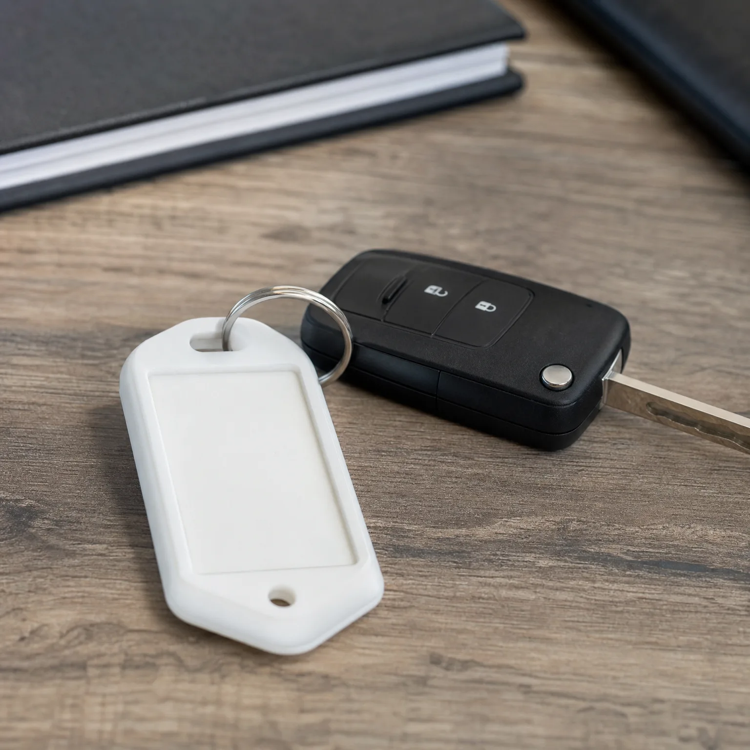 A black flip-style car key with two buttons and a blank white plastic key tag on a split ring, lying on a wood-grain surface near a dark notebook.