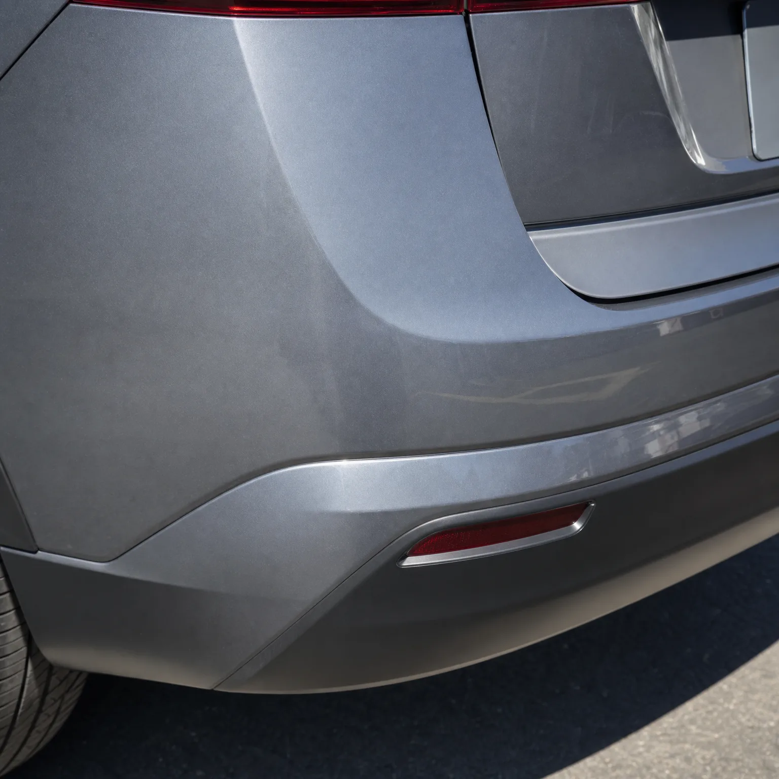 Close-up of the rear bumper and lower tailgate corner of a gray car, showing a red reflector and part of a tire above dark pavement.