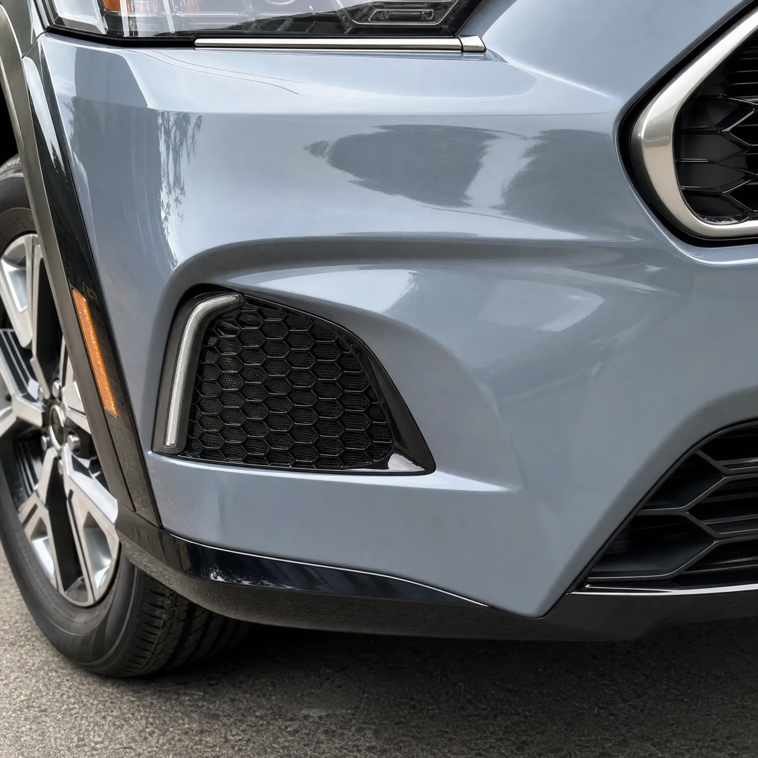 Close-up of the front left corner of a gray vehicle, showing the bumper with a black honeycomb mesh vent, chrome trim, part of the headlight and grille, and the front wheel over asphalt.
