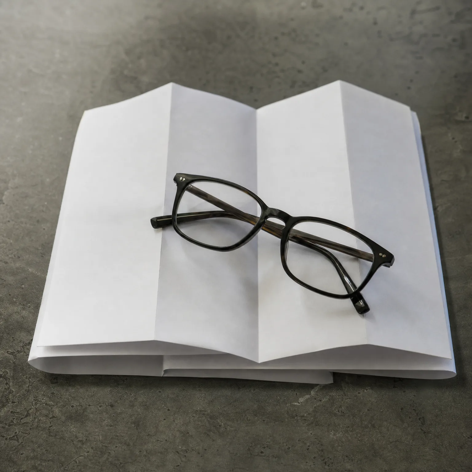 A pair of dark-framed eyeglasses rests on an open book with blank white pages, placed on a gray concrete surface.