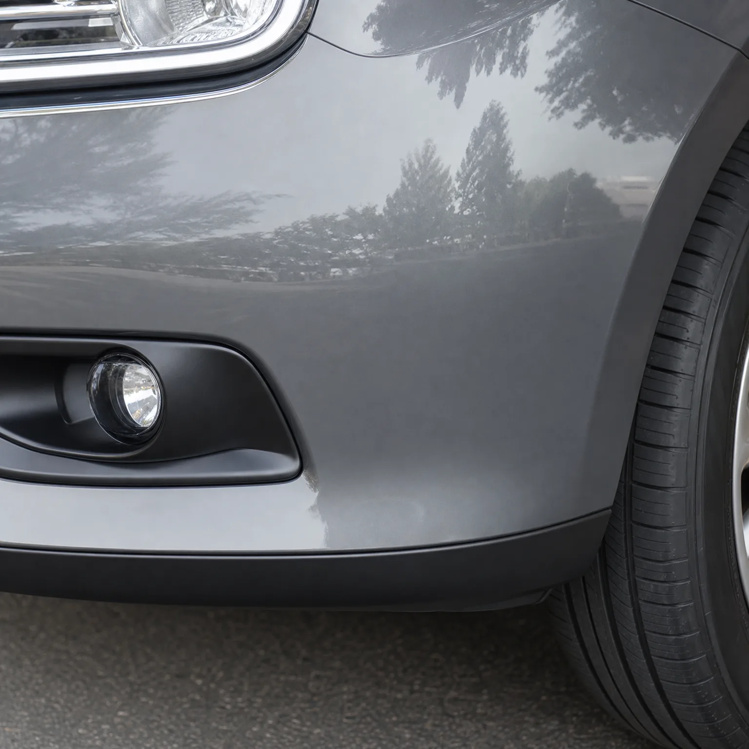 Close-up of the front corner of a gray car, showing the fog light in a black bumper surround, the front wheel with tire and alloy rim, and trees reflected in the glossy paint.