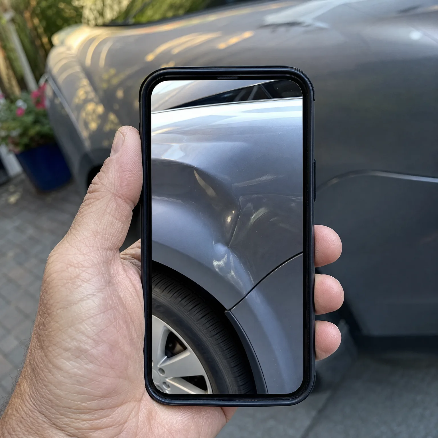 A hand holds a smartphone displaying a close-up photo of a dented gray car fender and wheel, in front of the same gray car parked on paving stones.