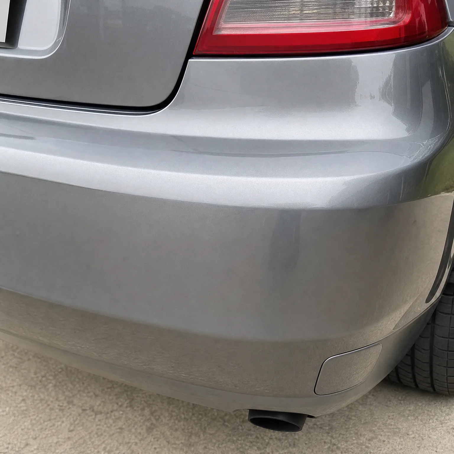 Close-up of the rear corner of a silver-grey car, showing the bumper, part of a red taillight, an exhaust tip, and a tyre, with pale pavement in the background.