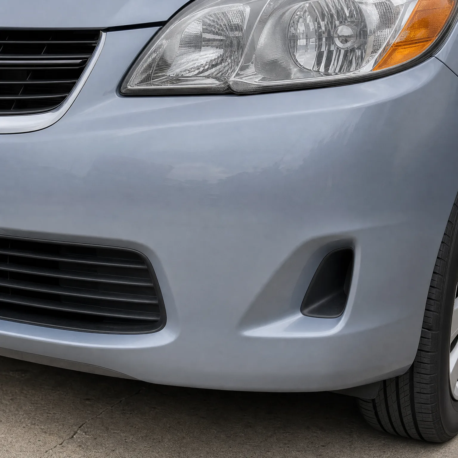 Close-up of the front left corner of a light blue-gray car, showing the headlight, bumper with lower grille and fog light opening, and part of the front tire on pavement.