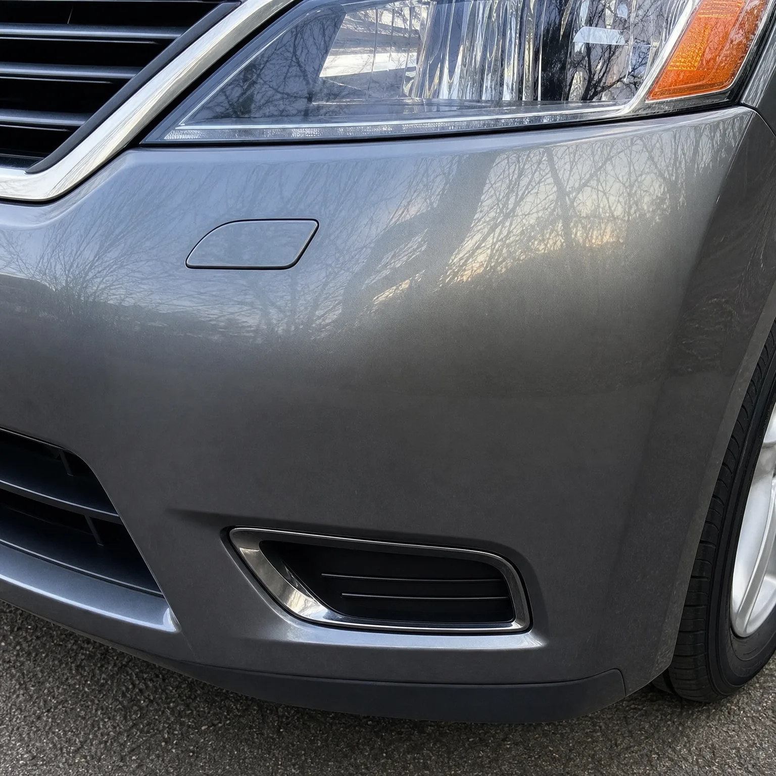 Close-up of the front left corner of a gray car, showing the headlight, fog light bezel, bumper and part of the tire and alloy wheel above asphalt.