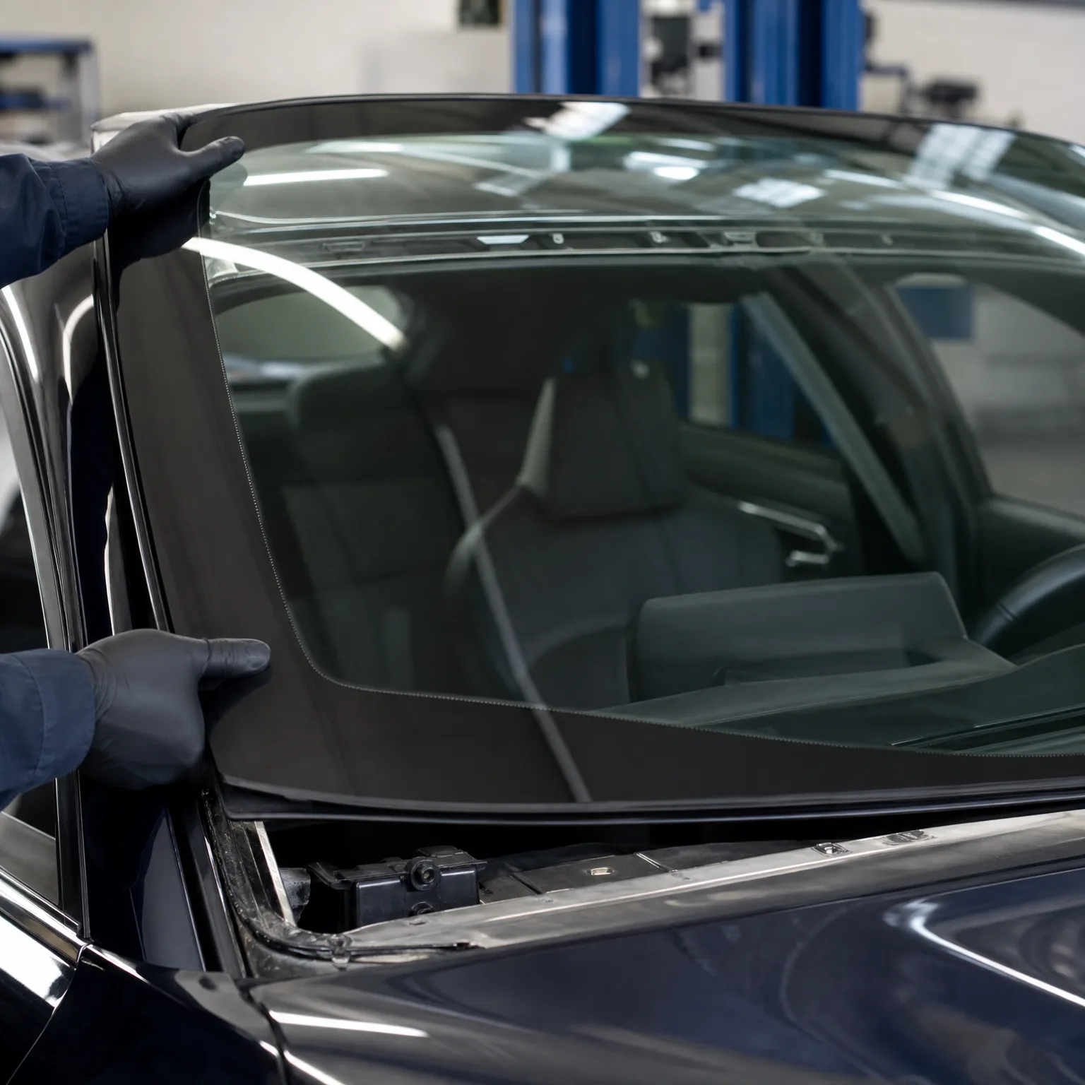 Gloved hands hold a windshield above the open frame of a dark car inside a workshop.