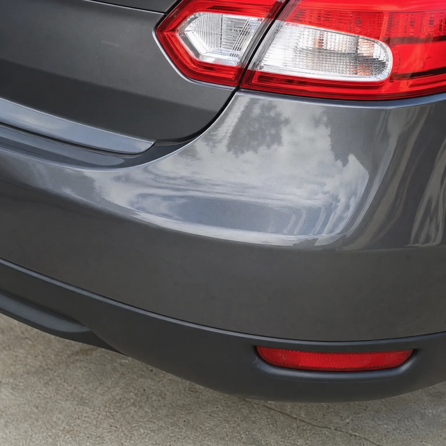 Close-up of the rear corner of a gray car showing a dent in the quarter panel above the bumper, with the red and clear taillight and a red bumper reflector visible, parked on concrete.