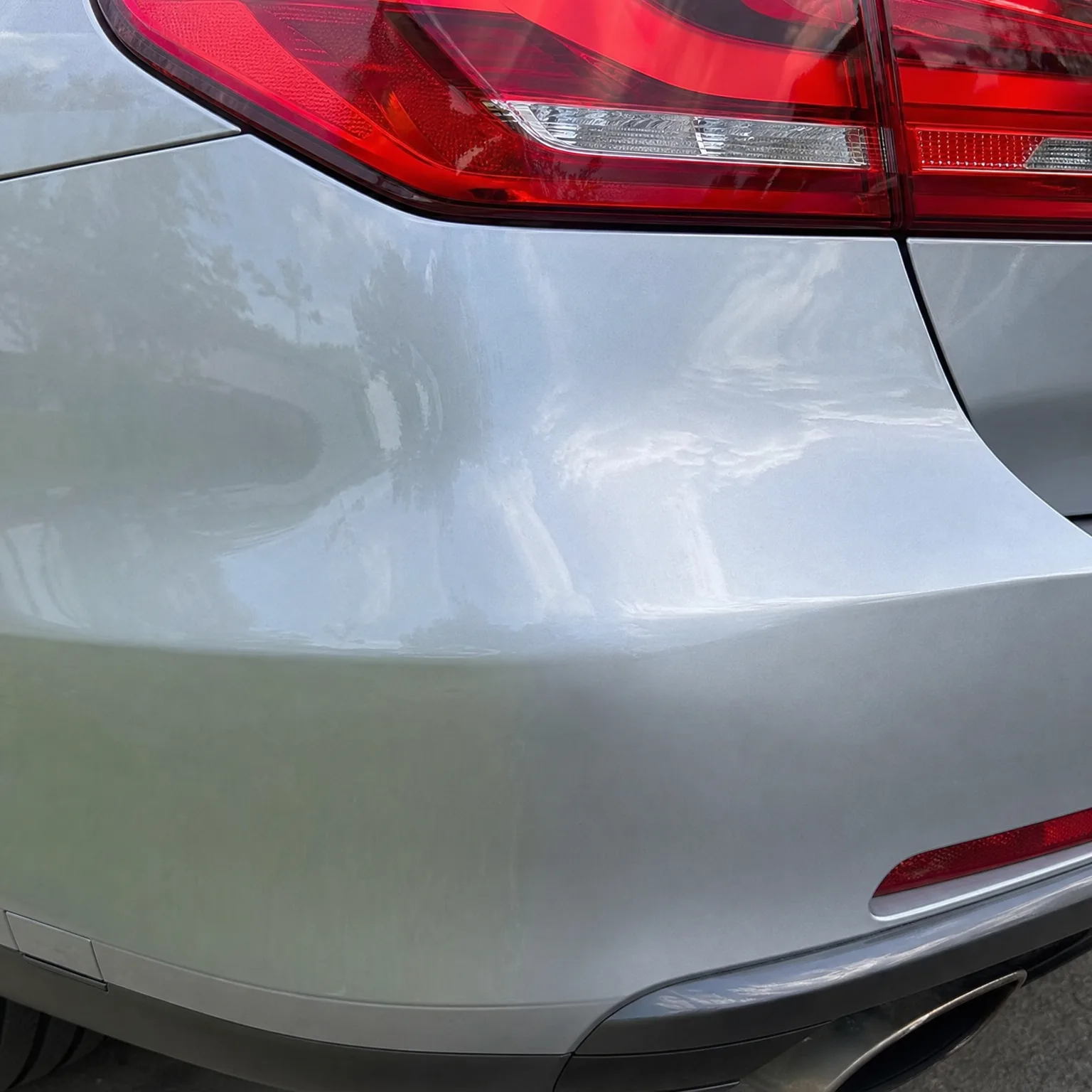Close-up of the rear corner of a silver car showing a red tail light, a dented and creased quarter panel and bumper, with tree reflections in the paint.