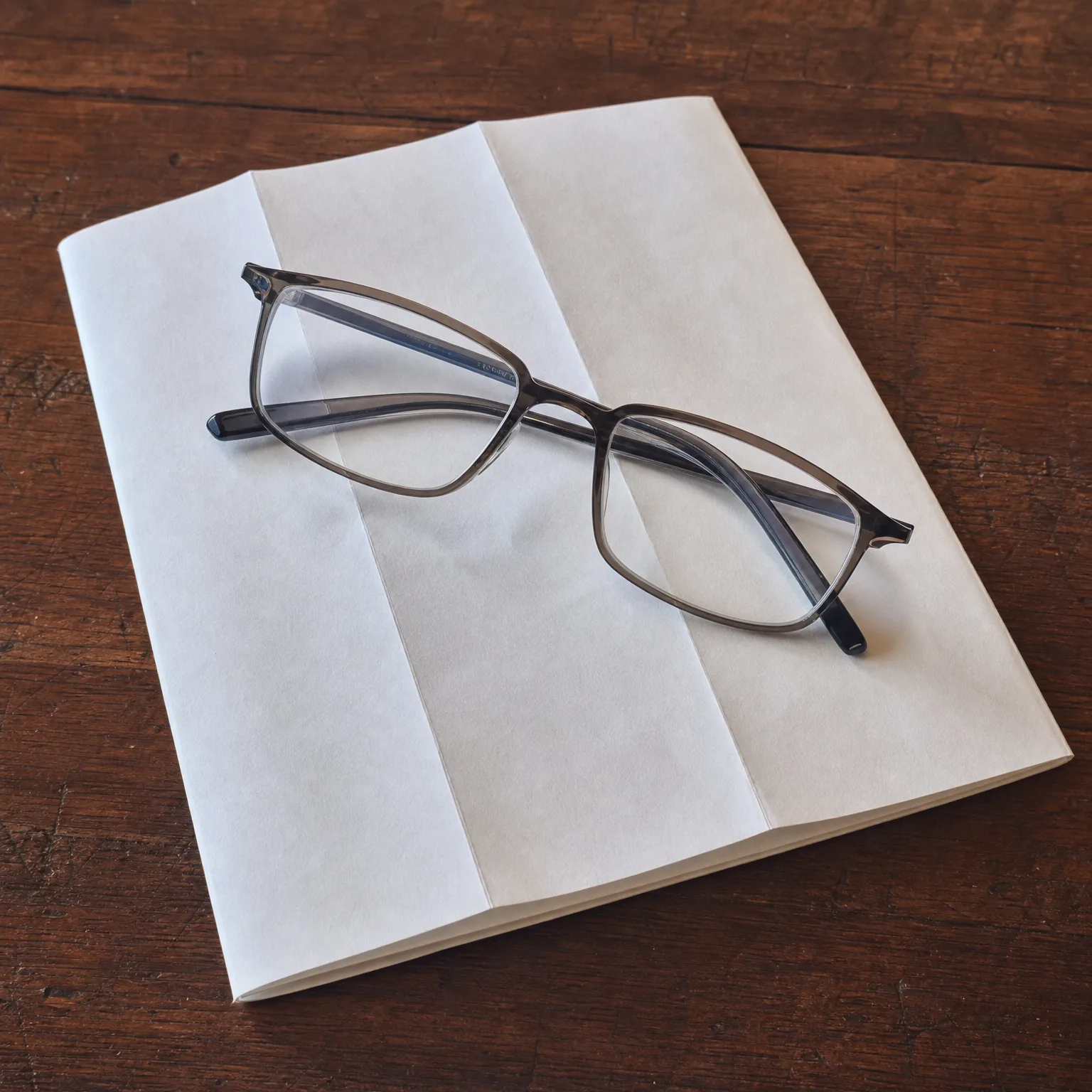 A pair of gray-framed eyeglasses resting on an open blank-paged booklet on a dark wooden table.