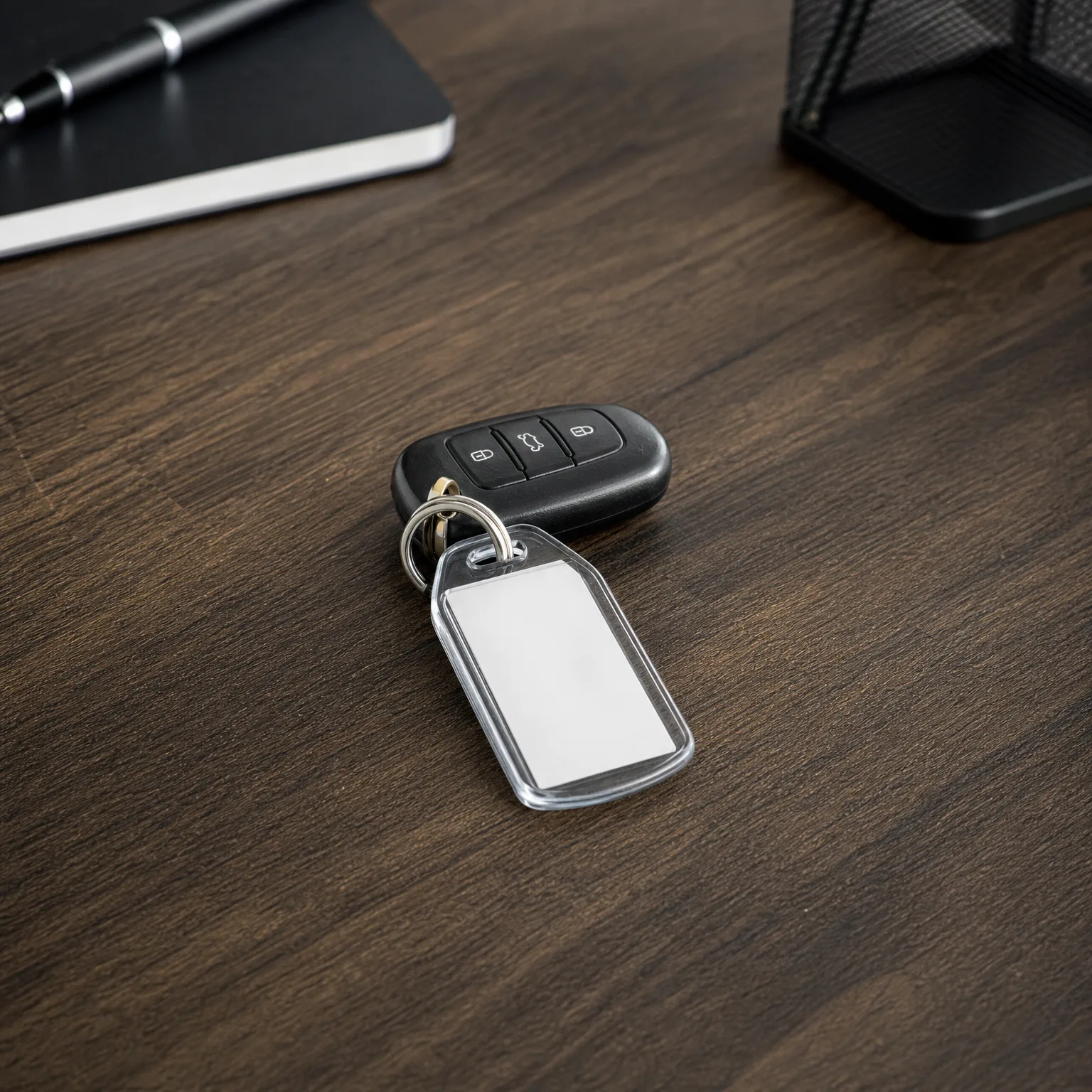 A black three-button car key fob with a blank white plastic keychain tag on a split ring, lying on a dark wood desk near a notebook with pen and a mesh desk organizer.
