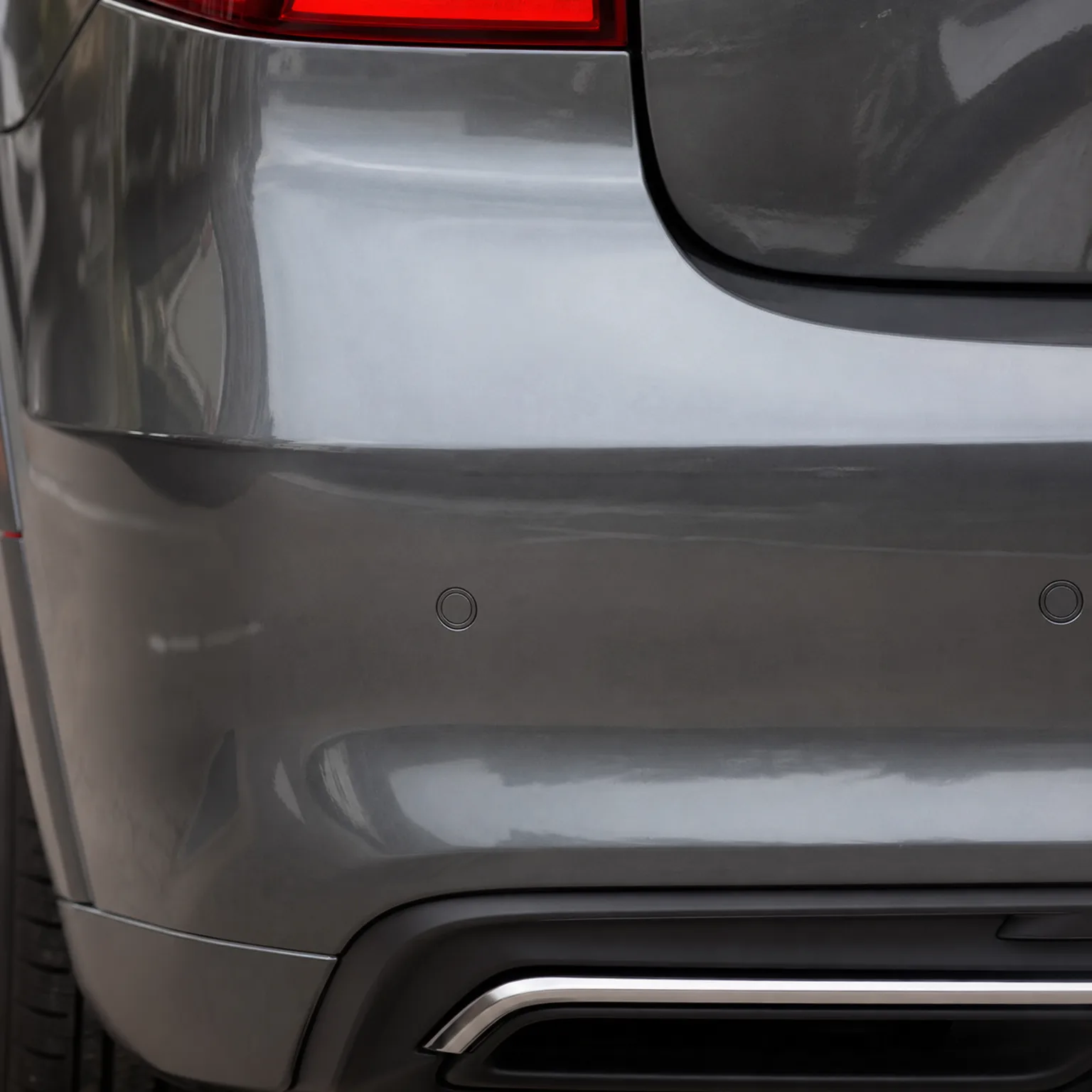 Close-up of the rear corner of a gray car showing the bumper with two parking sensors, part of a red taillight, a chrome trim strip low on the bumper, and a tire at lower left.