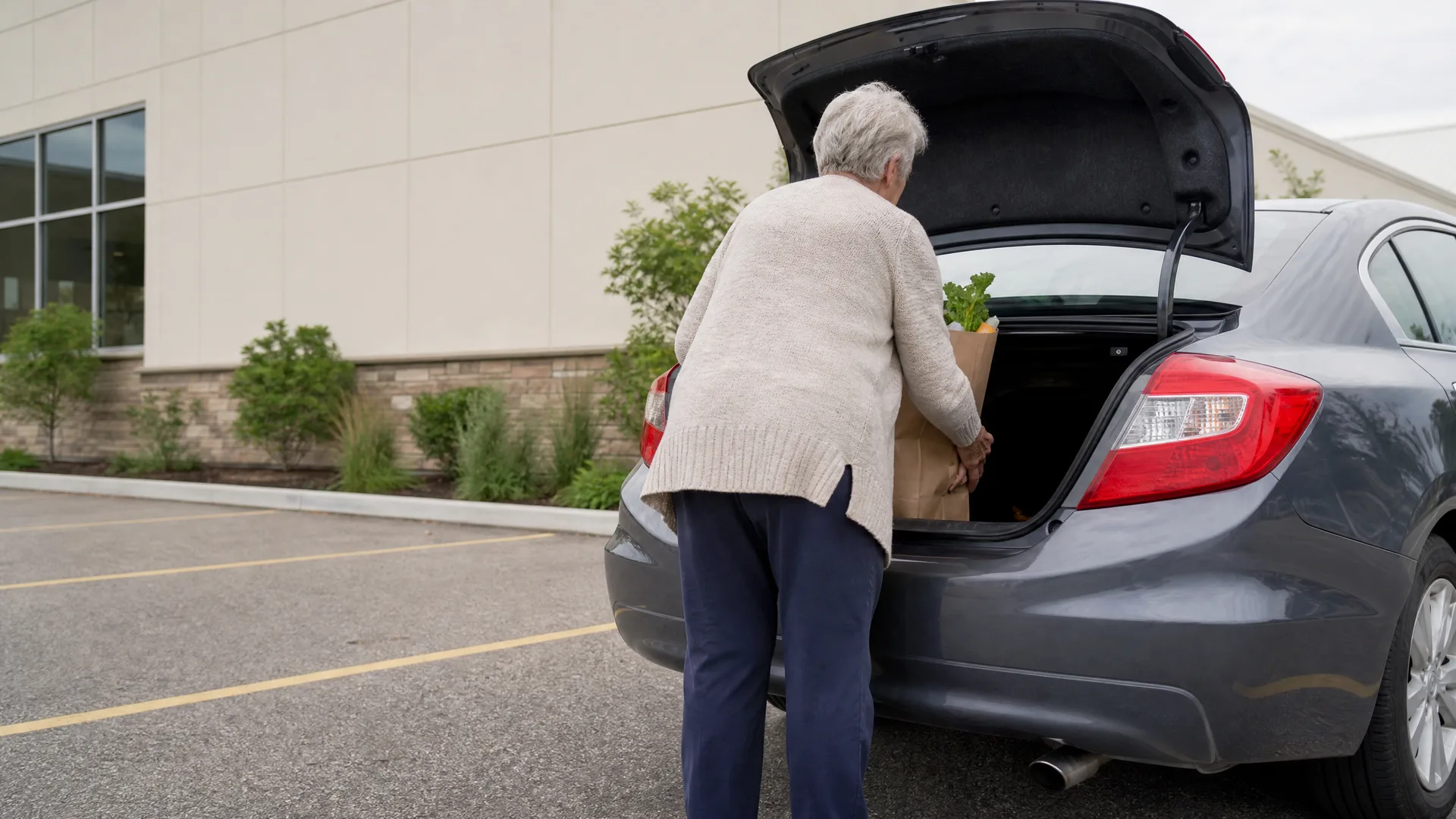 An older person with white hair in a cream cardigan and dark trousers loads a paper grocery bag holding greens into the open trunk of a gray sedan in a parking lot beside a light-colored building.