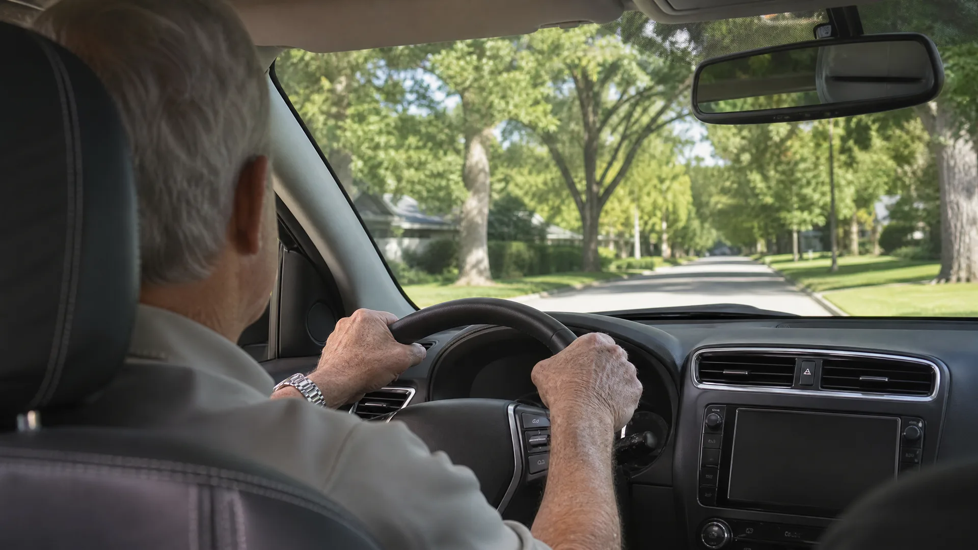 View from the back seat of a car showing an older man with gray hair holding the steering wheel while driving along a tree-lined street with houses and green lawns ahead.