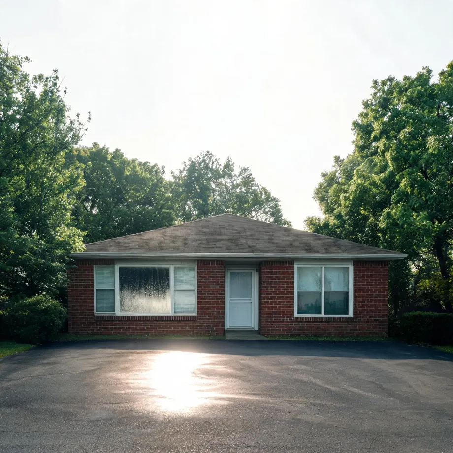 A single-story red brick building with a hipped shingle roof, a white door and white-framed windows, fronted by a large asphalt parking area with sun glare, surrounded by green deciduous trees under a bright hazy sky.