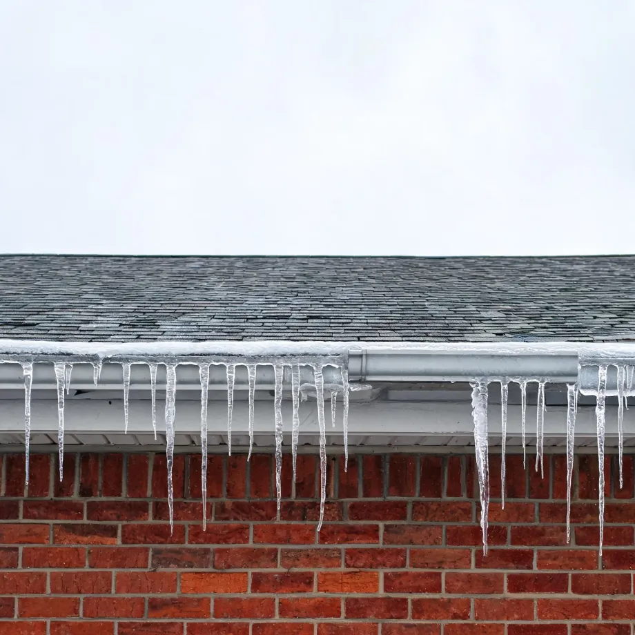 Icicles hang from a white gutter along the edge of a gray shingled roof above a red brick wall, under an overcast white sky.