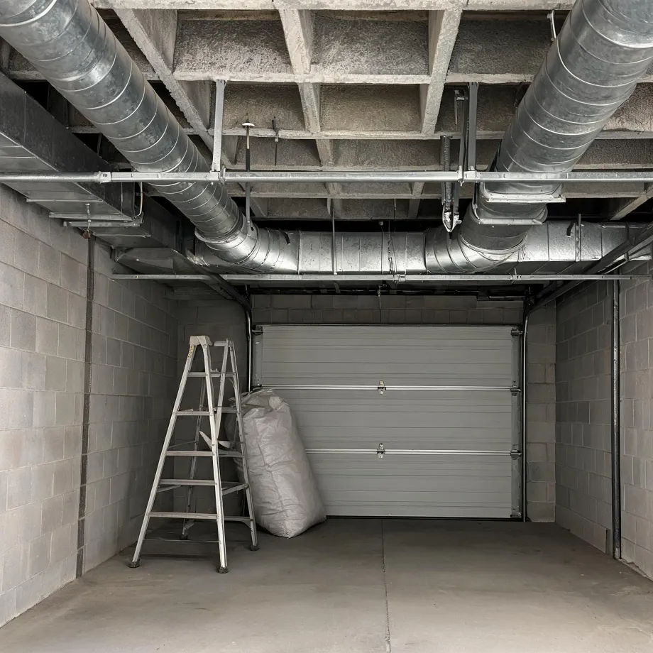 Interior of an empty concrete-block garage with a closed sectional roller door, exposed metal ventilation ducts under a coffered concrete ceiling, an aluminium step ladder and a large white bag on the floor.