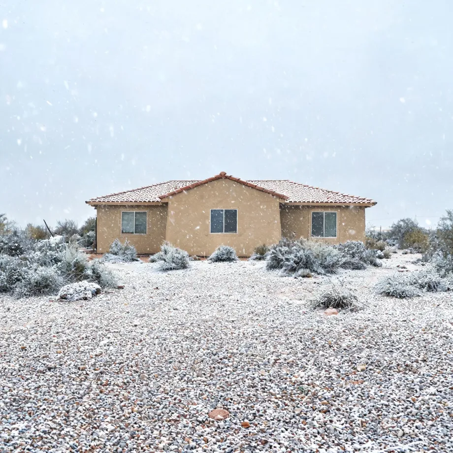 A single-story tan stucco house with a tile roof stands behind snow-dusted desert shrubs and a gravel yard during falling snow.