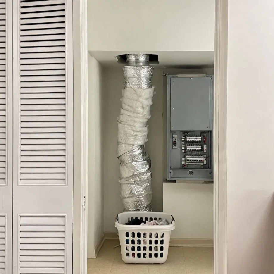 A narrow interior closet with a flexible foil dryer duct running from the floor to a ceiling vent, wrapped in loose white insulation, beside an open electrical breaker panel, with a white laundry basket of clothes on the tiled floor and louvered doors at left.