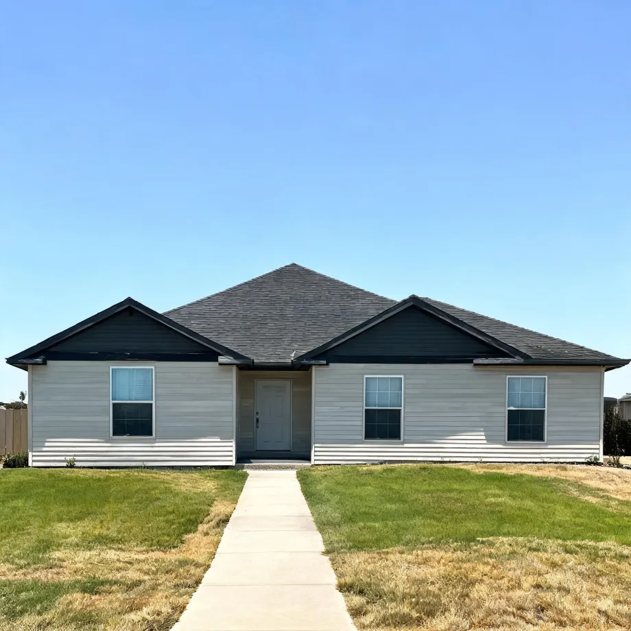Single-story house with light gray horizontal siding, dark gray shingled roof with two front gables, a white front door, and three windows, fronted by a concrete walkway and patchy green and brown lawn under a clear blue sky.
