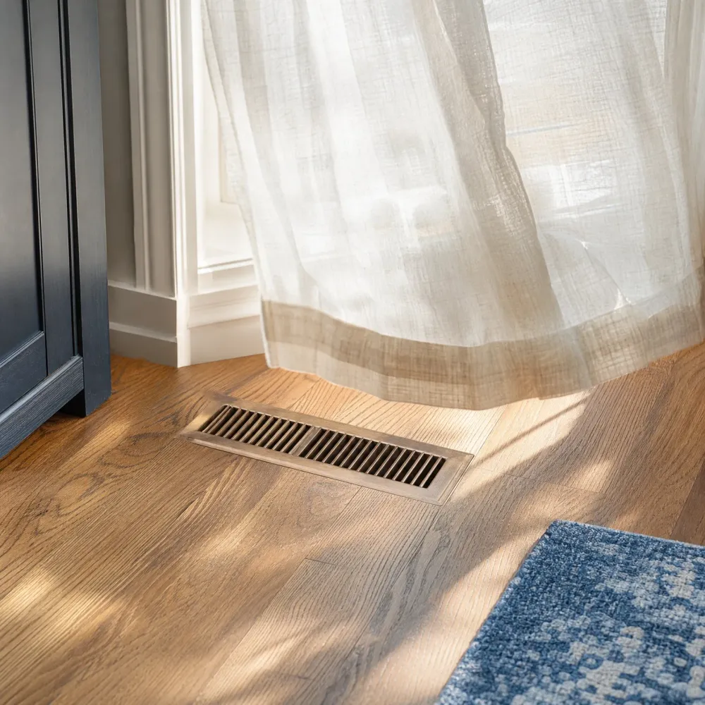 A rectangular metal floor vent set into a hardwood floor near a white baseboard and dark cabinet, with a sheer curtain hanging above and a blue patterned rug at the lower right.