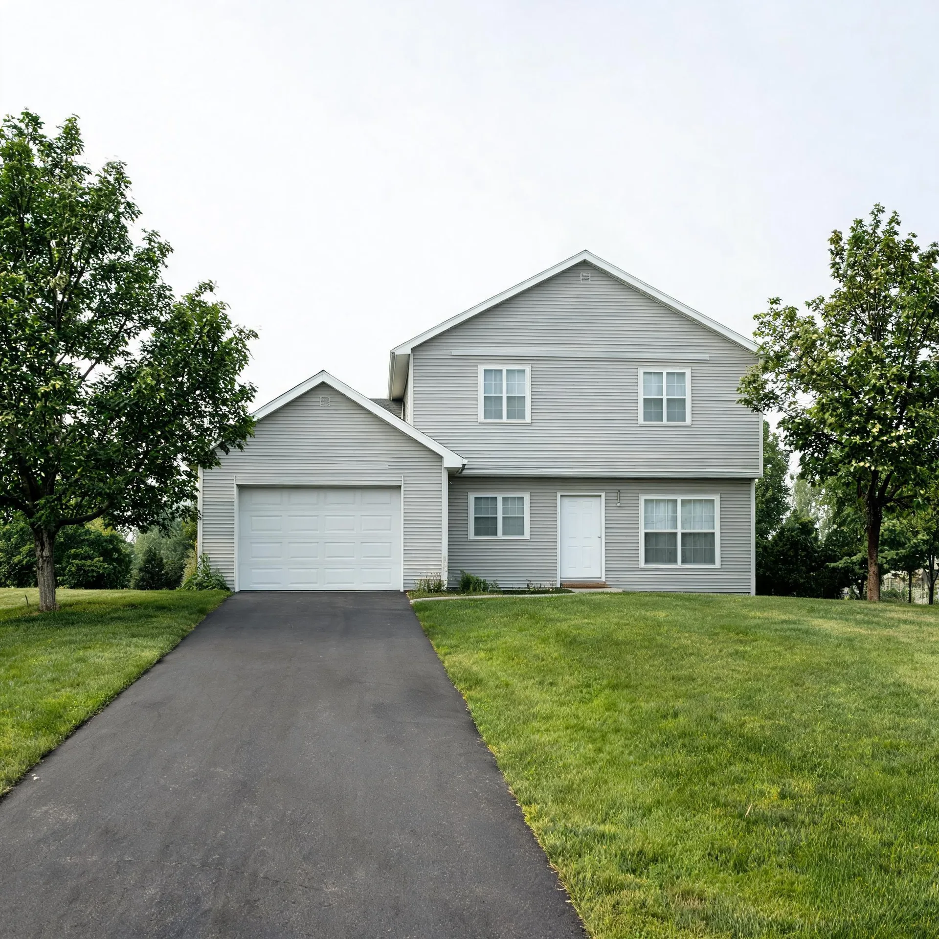 A two-story light gray vinyl-sided house with an attached single-car garage, a paved driveway leading to it, mown grass lawn, and deciduous trees on both sides under an overcast sky.
