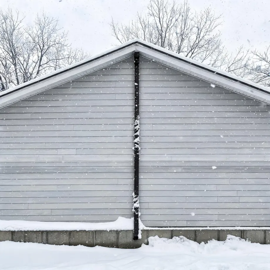 Gable end of a gray horizontal-sided building with a dark downspout running down the center to a concrete foundation, with snow on the ground and falling, and bare trees behind the roofline.