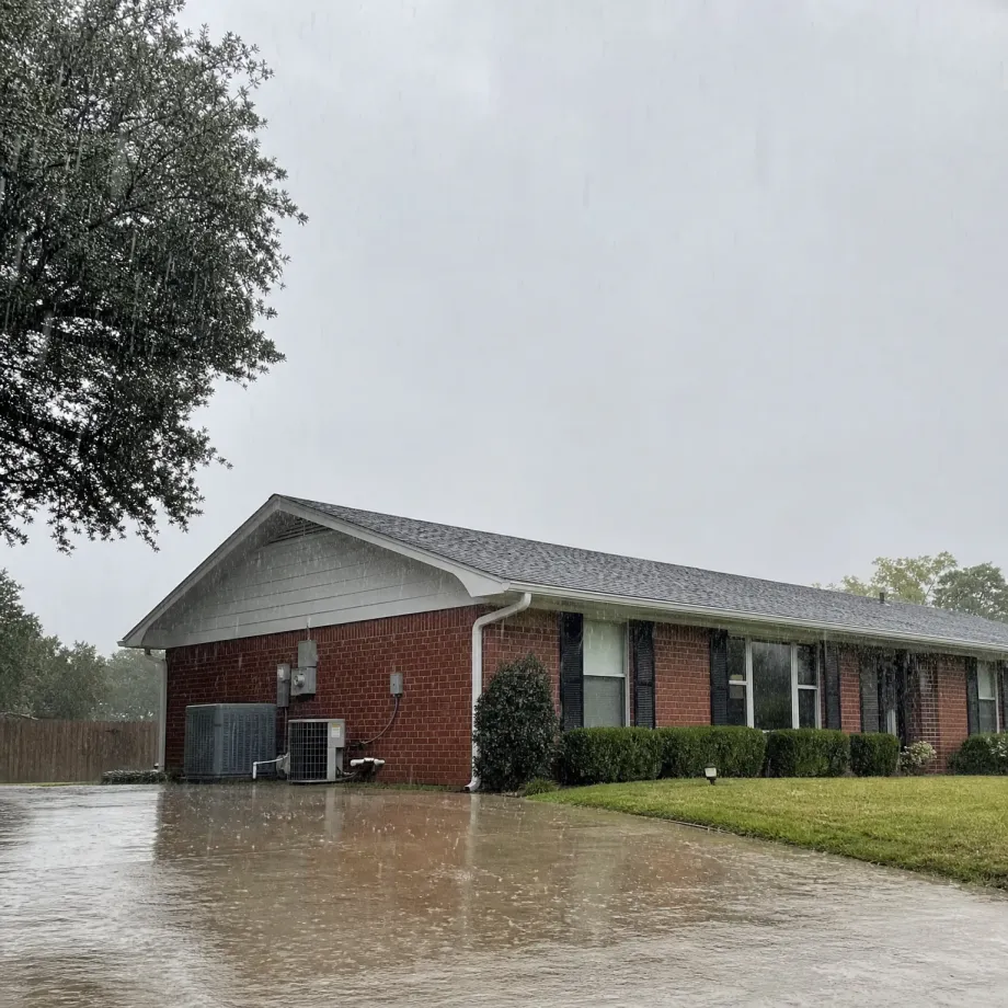 Heavy rain falls on a single-story red brick house with a gray shingled roof, air conditioning units along the side wall, and a flooded driveway in the foreground under an overcast sky.