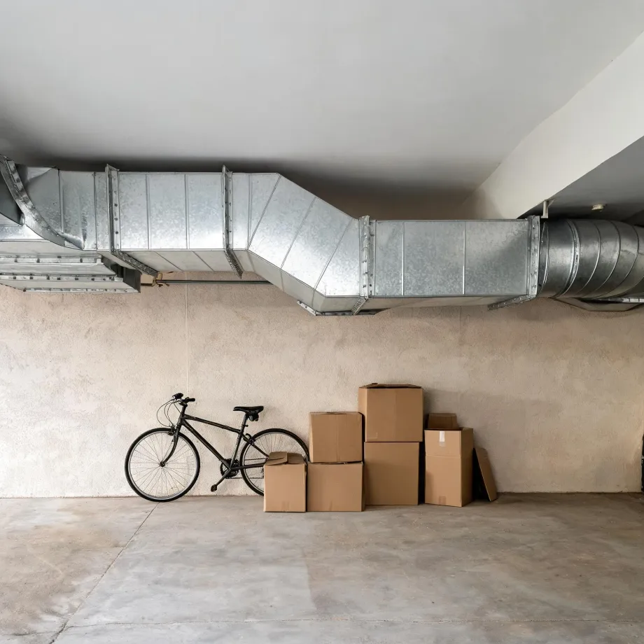 Interior basement or garage space with large galvanized sheet-metal ductwork running along the ceiling, a bicycle leaning against a plaster wall, and several stacked cardboard boxes on a concrete floor.
