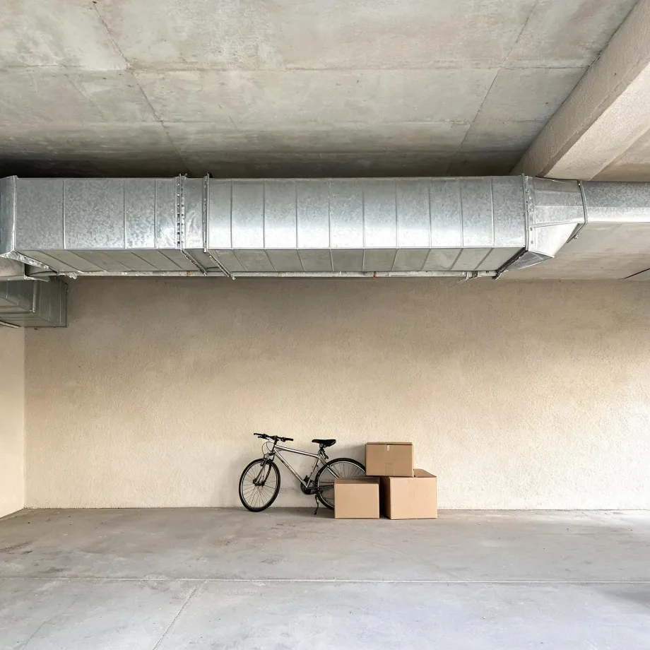 Empty concrete-floored interior space with a large galvanized ductwork running below the concrete ceiling, and a bicycle leaning against a beige stucco wall next to three cardboard boxes.