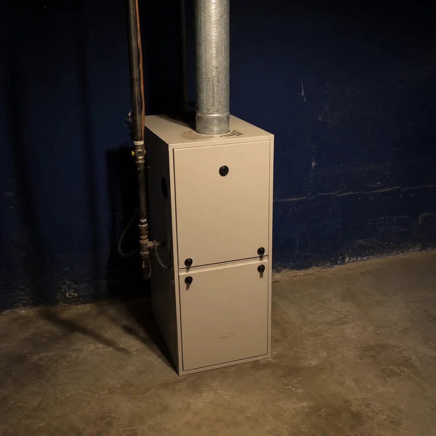 A beige furnace with a metal flue pipe stands against a dark blue painted basement wall on a concrete floor, with pipes running up the wall beside it.