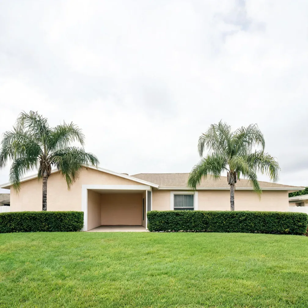 A single-story beige stucco house with a shingled roof and open carport, flanked by four palm trees and a trimmed hedge, with a mown lawn in front under an overcast sky.