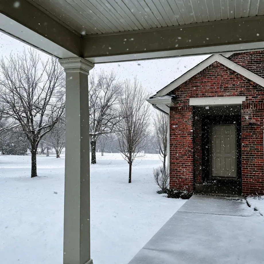 View from under a covered porch with a white column toward a red brick house entrance with a gray door, with falling snow, snow-covered ground and bare trees.