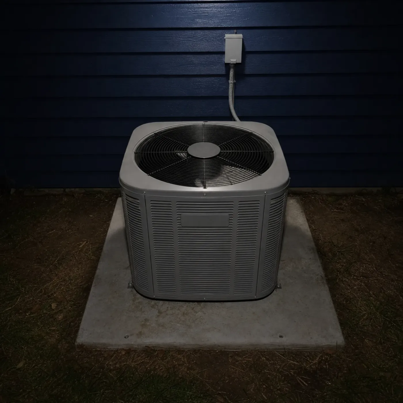 A gray outdoor condenser unit with a top-mounted fan grille sits on a concrete pad against dark blue lap siding, with a conduit running to a small wall-mounted disconnect box, photographed at night.